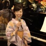 浴衣を着たシンガーソングライターがピアノの前でマイクを持ちながら演奏している様子