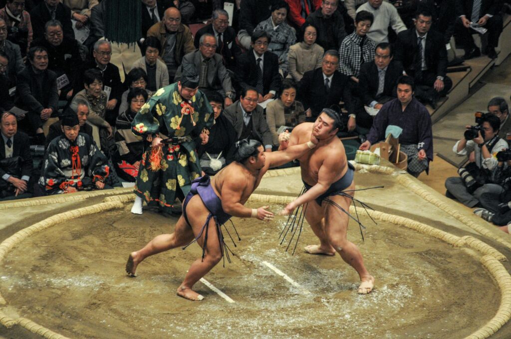 国技館の土俵で相撲を取る力士と行司、観客席には大勢の観客とカメラマンが見守る熱戦の様子