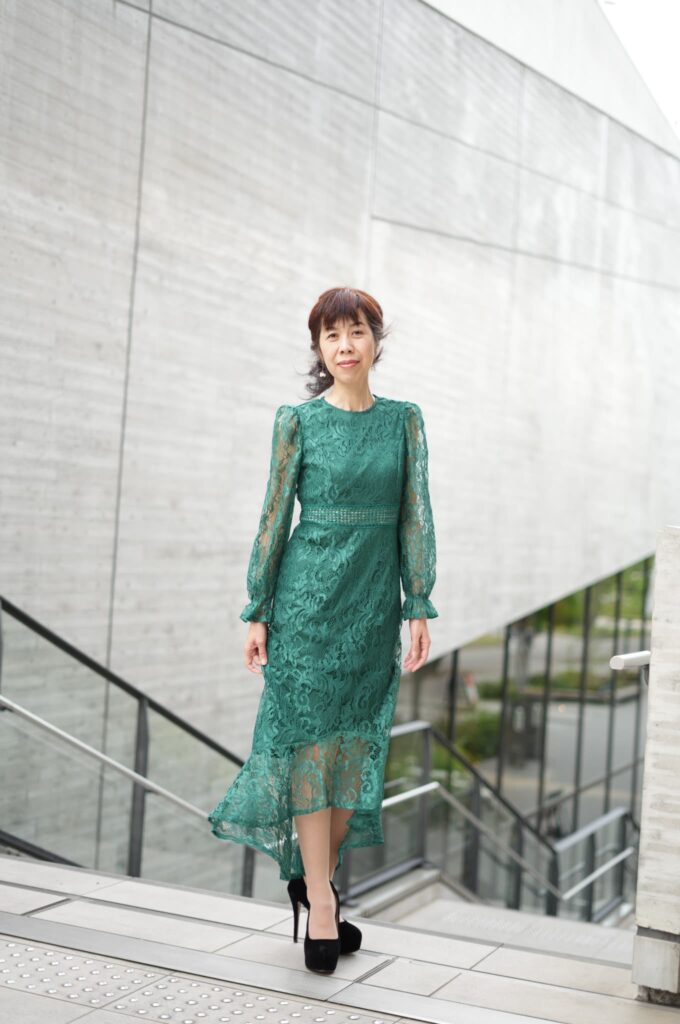 Ageless model in a noble green lace dress, standing naturally by a wall in harmony with the surrounding light.