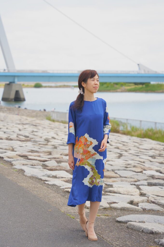 紺青地に川と波の幾何学模様の着物ドレスを着たモデルが川辺を歩く全身写真